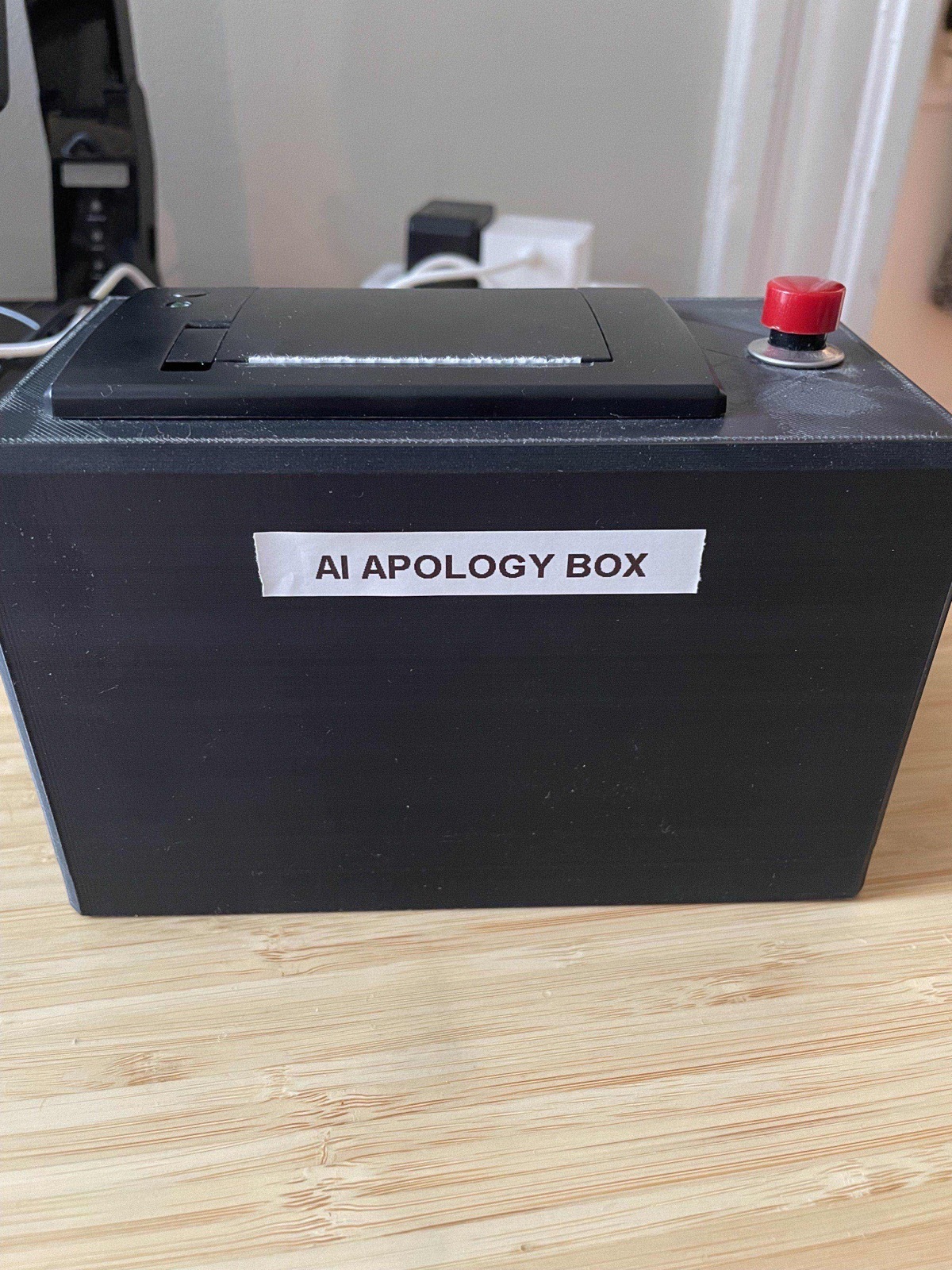 A black 3D-printed box labeled 'AI Apology Box' with a receipt printer on top and a red button