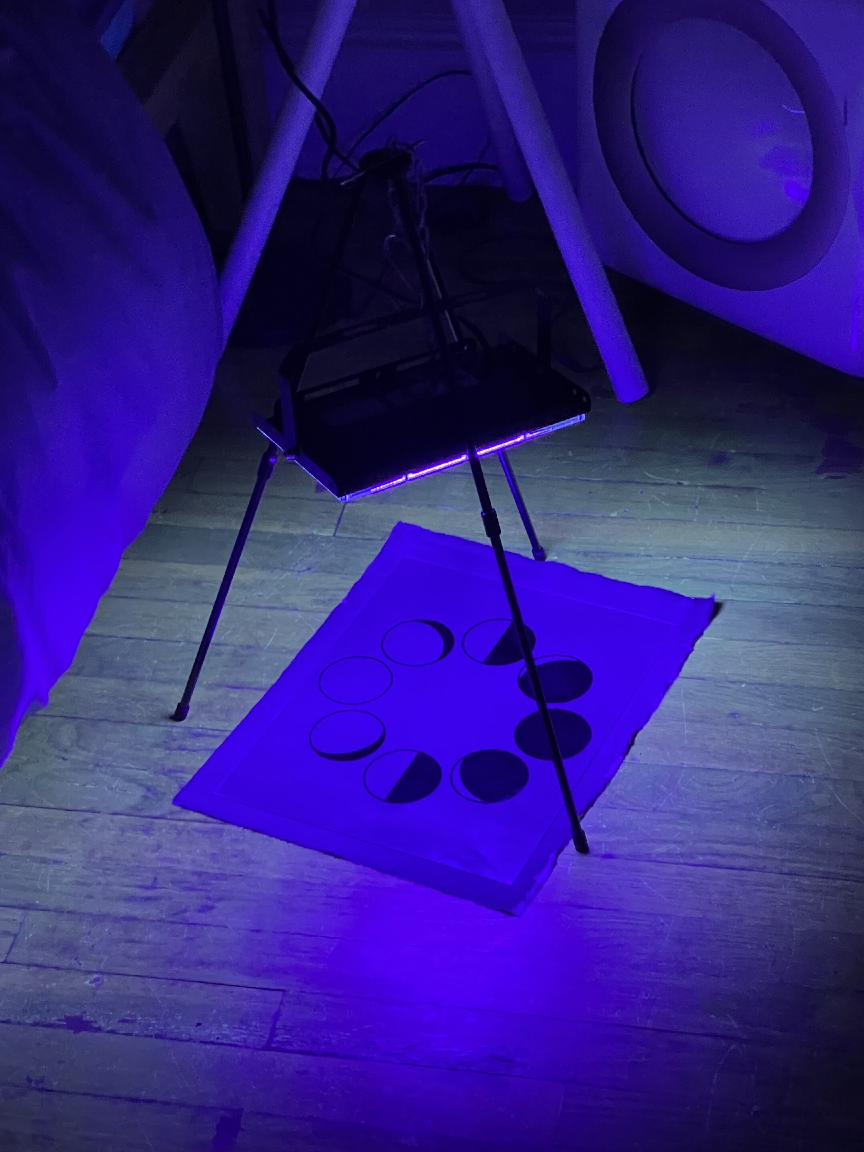 UV lamp on a tripod exposing a cyanotype print on the floor, bathed in purple light